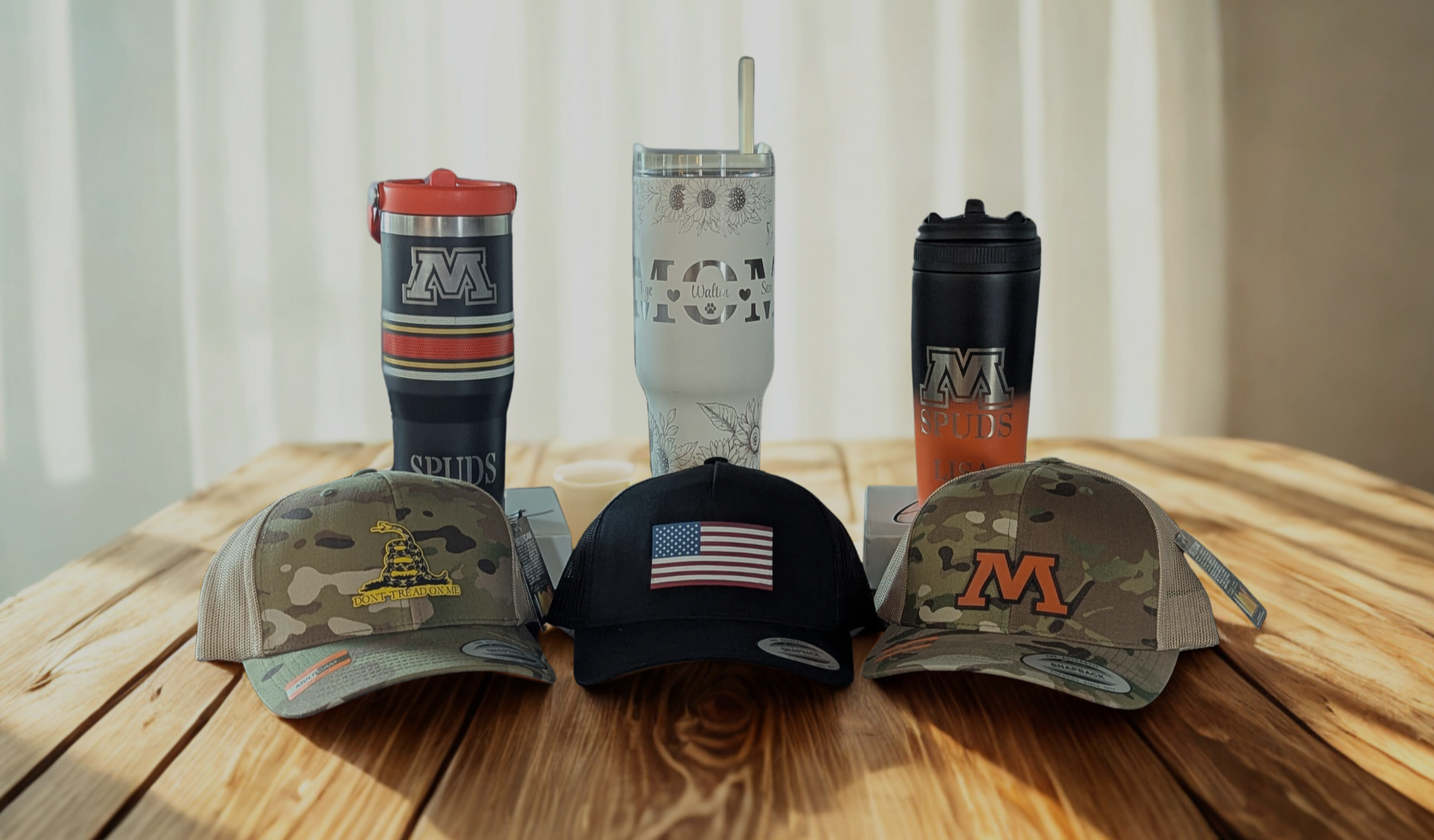 Three caps and three custom engraved tumblers on a wooden surface with a neutral background